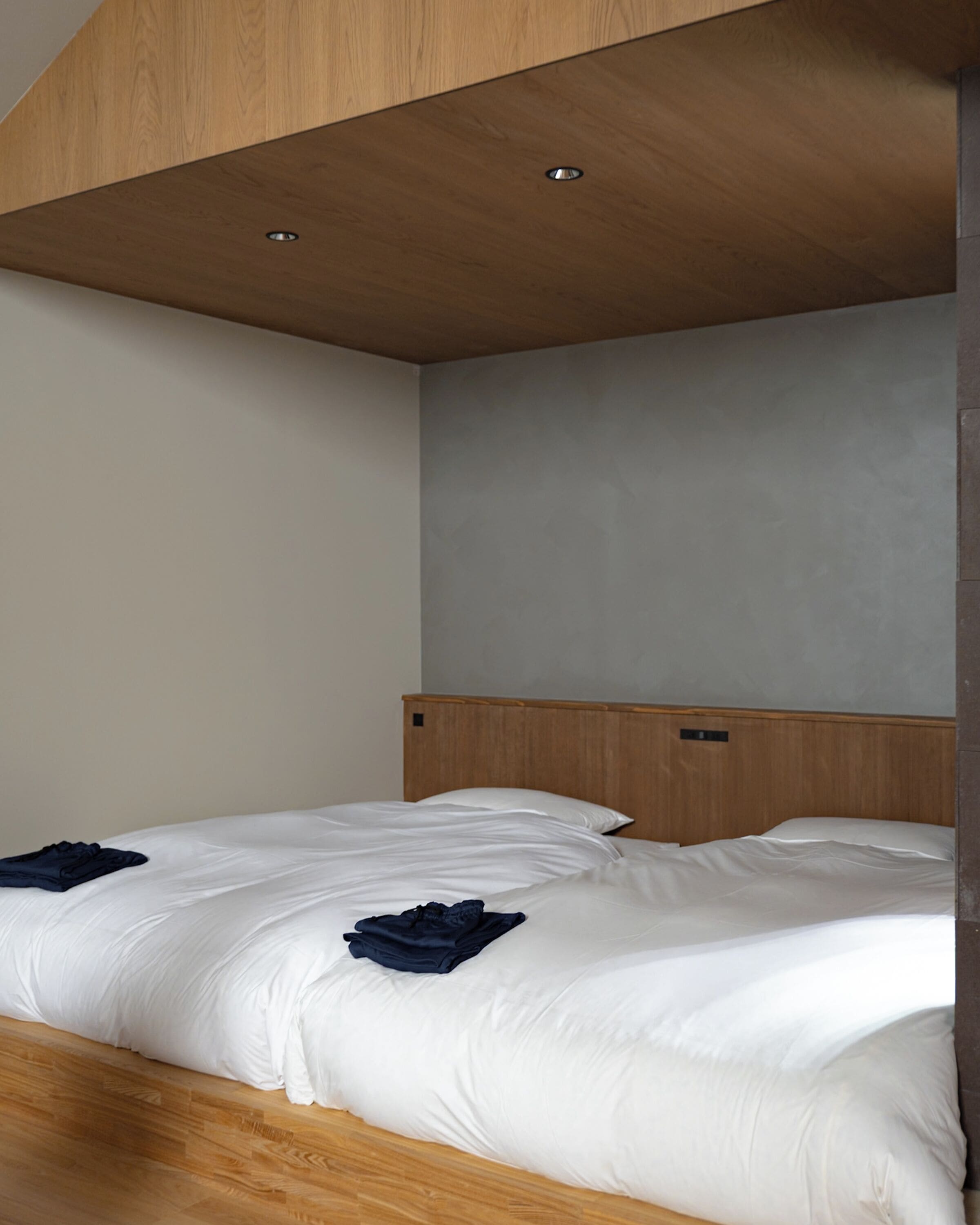 Ein Einzelbett mit Holz Kopfteil, eine graue Wand und eine Holzbalkendecke mit Einbaustrahlern.