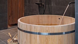 Una bañera de madera con cabezal de ducha y manguera, una puerta de madera y suelo de baldosas.