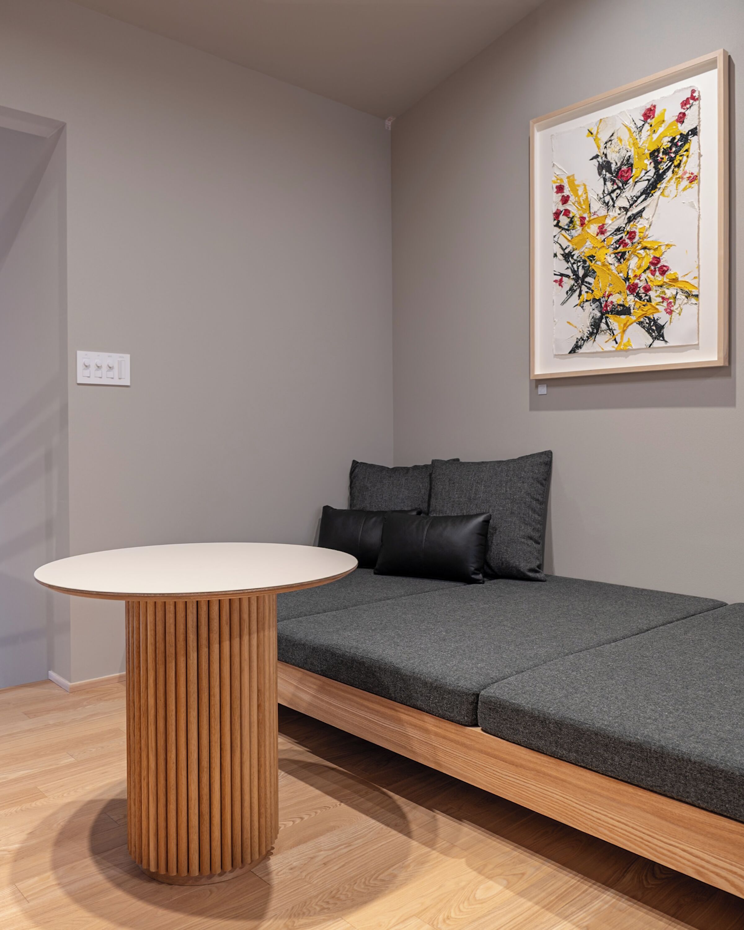Een kamer met een ronde houten tafel, een grijze bank met zwarte kussens en een lijst met een abstract schilderij aan de muur.