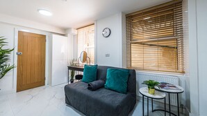 A modern living room with a grey sofa, a small round table, and a wooden door. - Elegant 1BR Heathrow Direct West Kensington (London)
