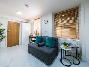A modern living room with a grey sofa, a small round table, and a wooden door. - Elegant 1BR Heathrow Direct West Kensington (London)