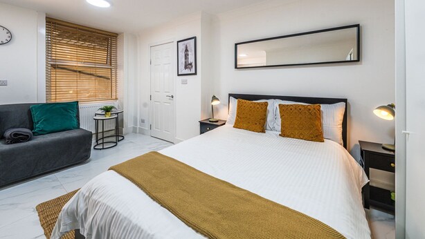 A modern bedroom with a bed, a sofa, a bedside table, and a mirror. - Elegant 1BR Heathrow Direct West Kensington (London)