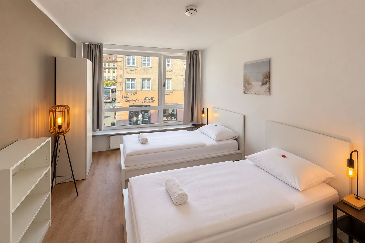 Habitación de hotel con dos camas, una ventana que ofrece vistas a edificios, un escritorio y una lámpara.