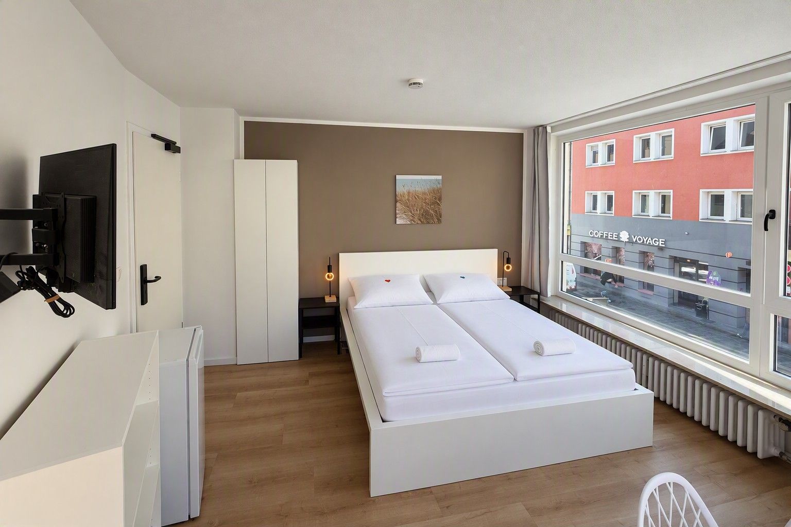 Une chambre d’hôtel moderne avec un grand lit, un téléviseur fixé au mur, une fenêtre donnant sur la ville et un radiateur placé sous la fenêtre.