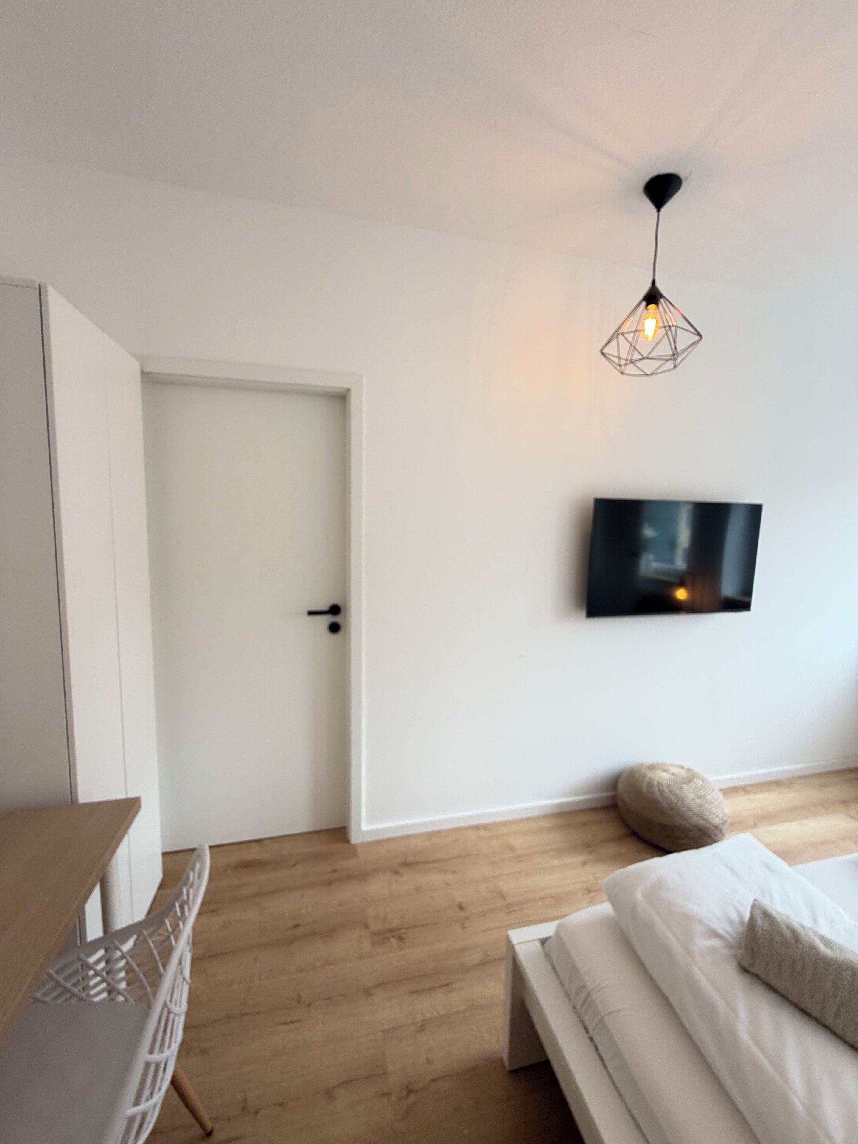 A bedroom with a bed, a TV mounted on the wall, a door, and a hanging light fixture.