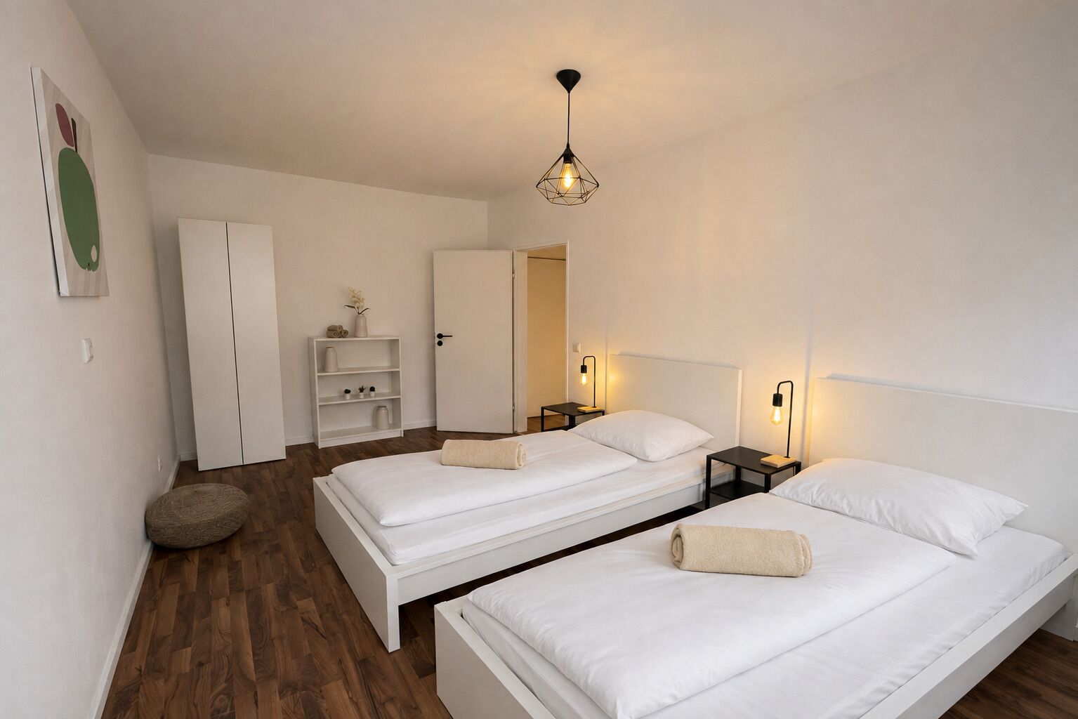 A modern hotel room with two single beds, a wardrobe, a small seating area, and a bedside table with a lamp.