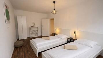 A modern hotel room with two single beds, a wardrobe, a small seating area, and a bedside table with a lamp.