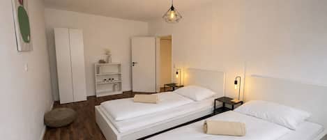 Quarto de hotel moderno com duas camas de solteiro, um guarda-roupa, um pequeno espaço de estar e uma mesa de cabeceira com abajur.