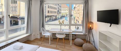 A hotel room with two beds, a dining table, chairs, a TV, and a view of the city.