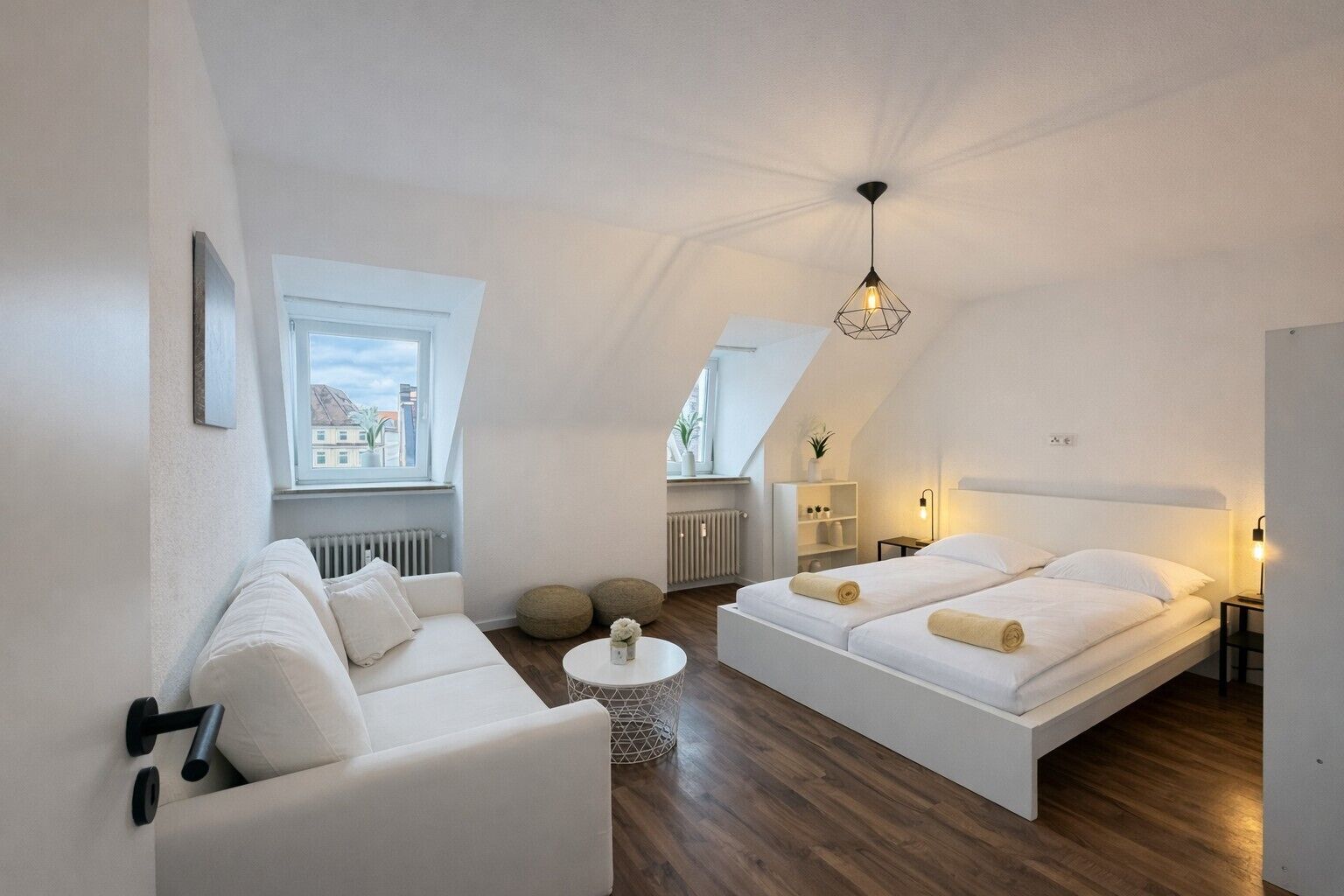 Une pièce moderne et bien éclairée, avec un canapé blanc, un lit avec deux oreillers, une petite table ronde et un luminaire suspendu.