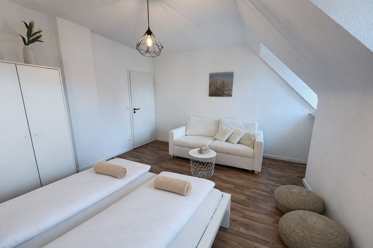 A modern, minimalist room with a white sofa, two single beds, a small round table, and a geometric pendant light.