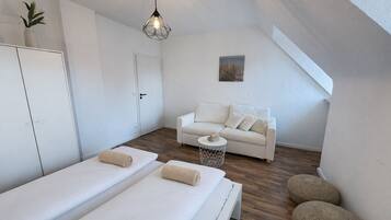 A modern, minimalist room with a white sofa, two single beds, a small round table, and a geometric pendant light.