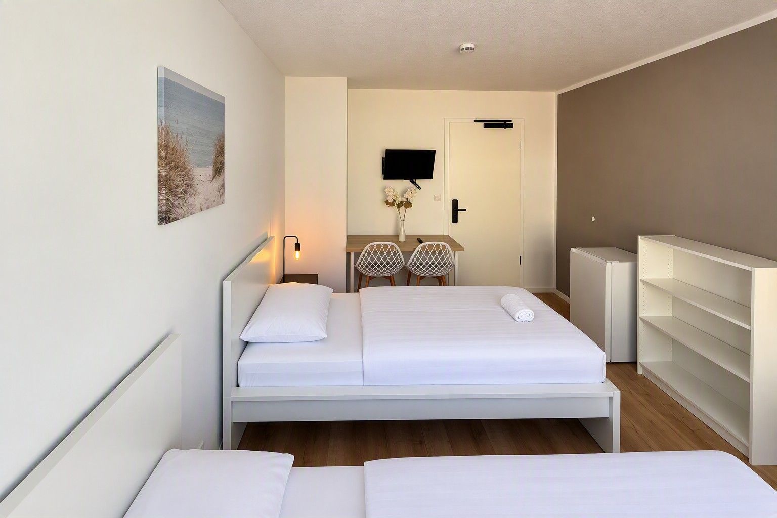A modern hotel room with a large bed, a desk, two chairs, a TV, and a bookshelf.