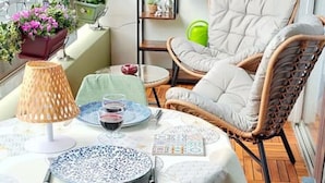Una terraza de madera con una mesa dispuesta para comer, rodeada de sillas de mimbre y un estante con plantas.