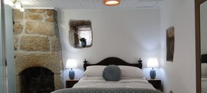 Une chambre avec un mur en pierre, une petite fenêtre ornée d’une plante, un lit avec deux lampes de chevet et une cheminée.