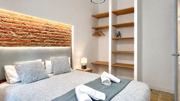 Une chambre moderne avec une tête de lit ornée d’un motif en briques, un lit avec une literie à motifs, un luminaire suspendu et des étagères en bois intégrées.