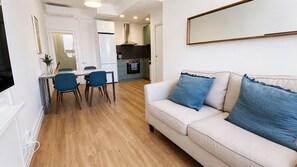 Ein Wohnzimmer mit einer Couch, zwei blauen Kissen, einem Essbereich mit Stühlen, einer Küche mit Schränken und einem an der Wand montierten Fernseher.