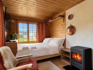 Ein gemütliches Schlafzimmer einer Blockhütte mit Bett, einem Holzstuhl, einem Kamin und einem Fenster mit Vorhängen.