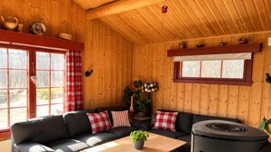 A cozy cabin interior with a sofa set, a wooden table, a stove, and a window with curtains.