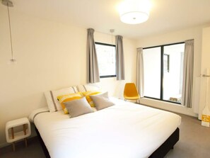 Ein modernes Hotelzimmer mit einem großen Bett, einem kleinen Beistelltisch, einem Sessel und einem großen Fenster mit Vorhängen.