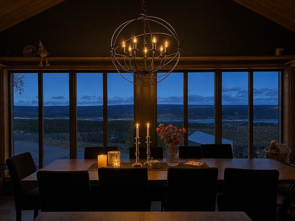 Comedor con una mesa grande, sillas, un candelabro, velas y vistas amplias a través de grandes ventanales.