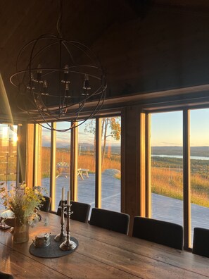 A dining area with a large table, chairs, a chandelier, and a view of a deck and landscape through large windows.