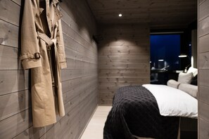 Quarto com paredes de madeira, uma cama com colcha preta e branca, e um casaco pendurado na parede.