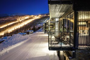Estrada coberta de neve que leva a um edifício com fachada de vidro, uma escadaria e vista para uma pista de esqui iluminada.