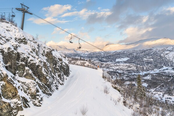 Trilha na montanha coberta de neve, com um teleférico e vista para um vale.