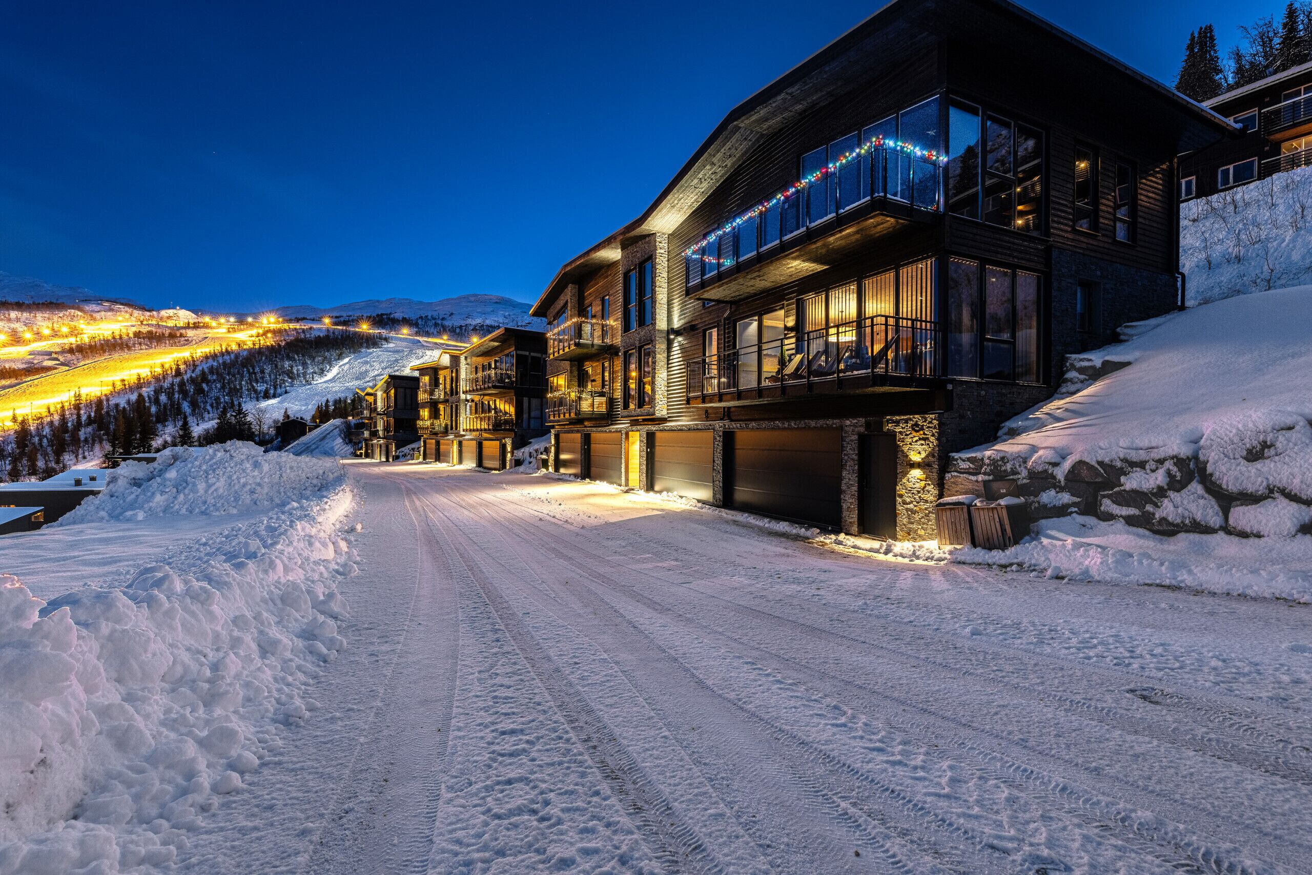 A snow-covered road leading to a modern building with large windows and a balcony.