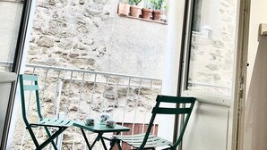 Ein Zimmer mit Glastür, die auf einen Balkon mit Tisch und Stühlen führt, sowie ein Fenster mit Blumentöpfen und eine Steinwand.