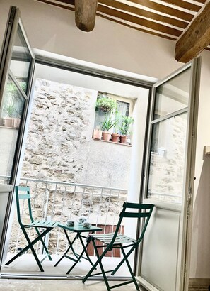 Een kamer met een glazen deur die leidt naar een balkon met een tafel en stoelen, een raam met potplanten en een stenen muur.