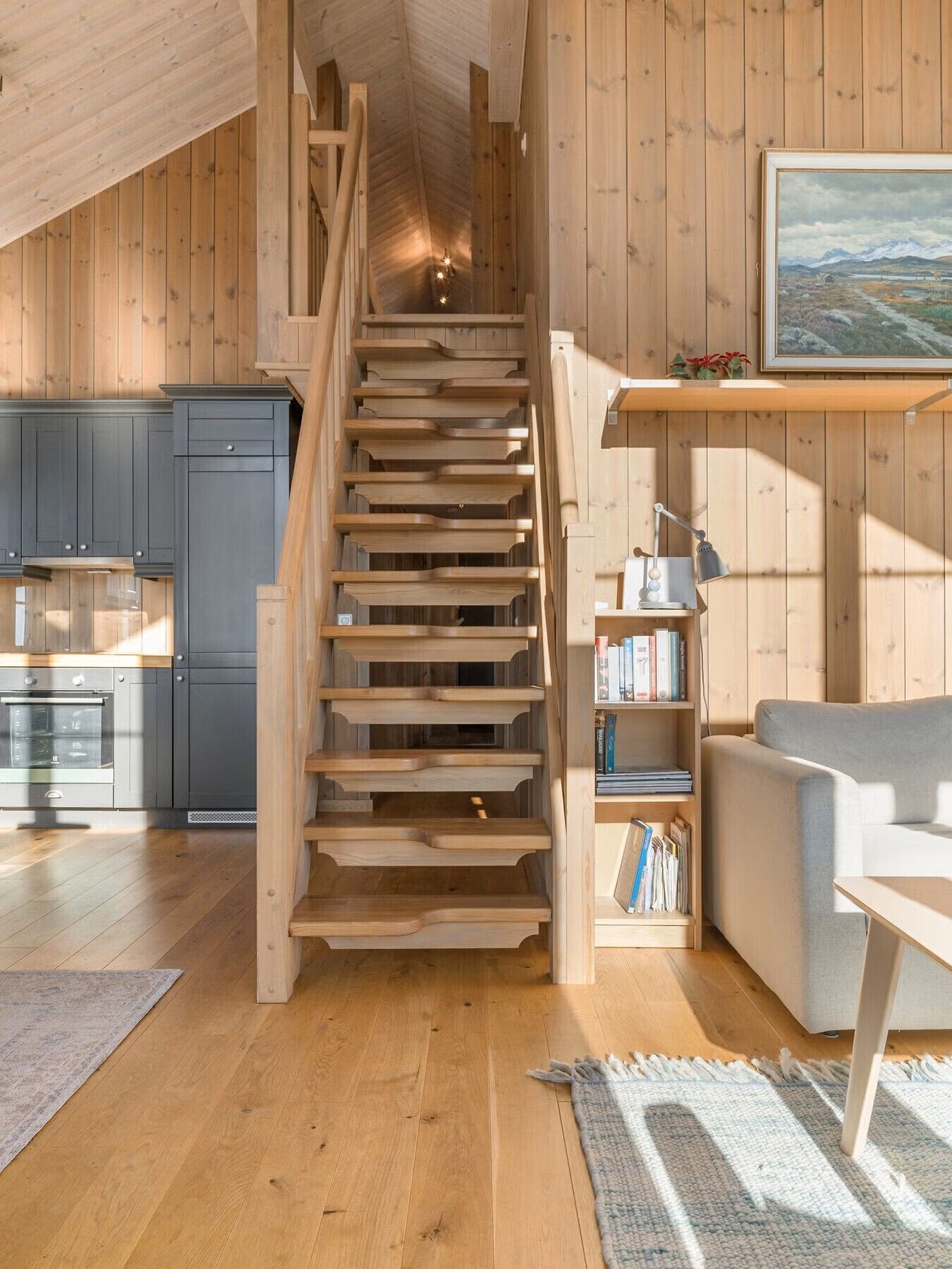 Un escalier en bois menant à une chambre aménagée en mezzanine, une cuisine avec des meubles sombres, une bibliothèque et un canapé gris.