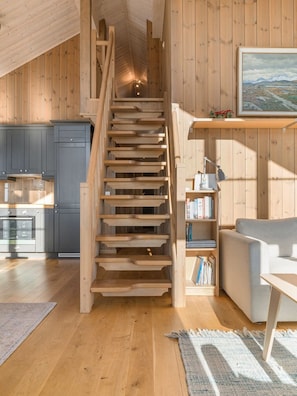 A wooden staircase leading to a loft bedroom, a kitchen with dark cabinets, a bookshelf, and a gray sofa.