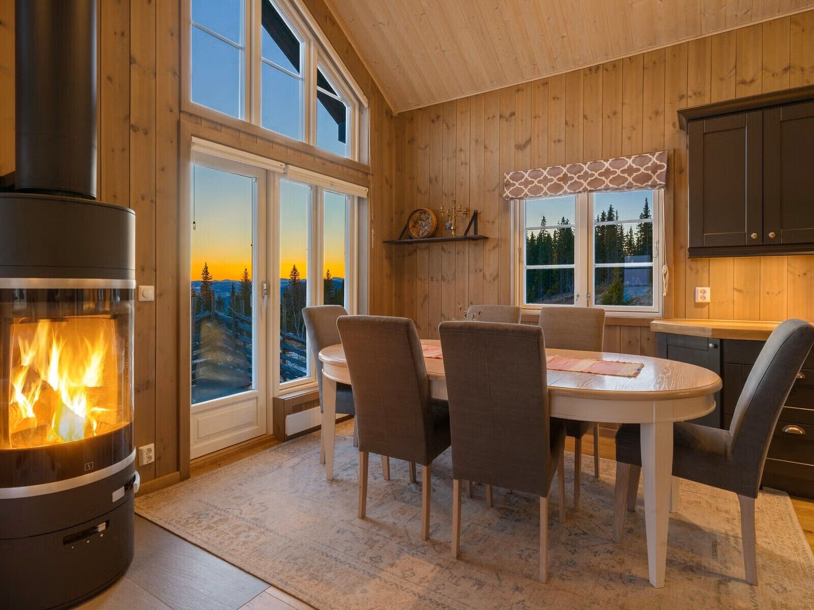 Interior acogedor de una cabaña con estufa de leña, una mesa de comedor con sillas y ventanas amplias que ofrecen vistas a un paisaje boscoso.