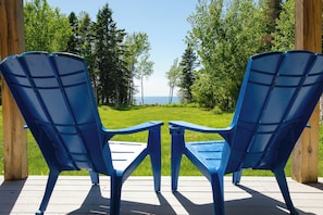Twee blauwe Adirondack-stoelen op een houten terras met uitzicht op een grasveld en bomen.