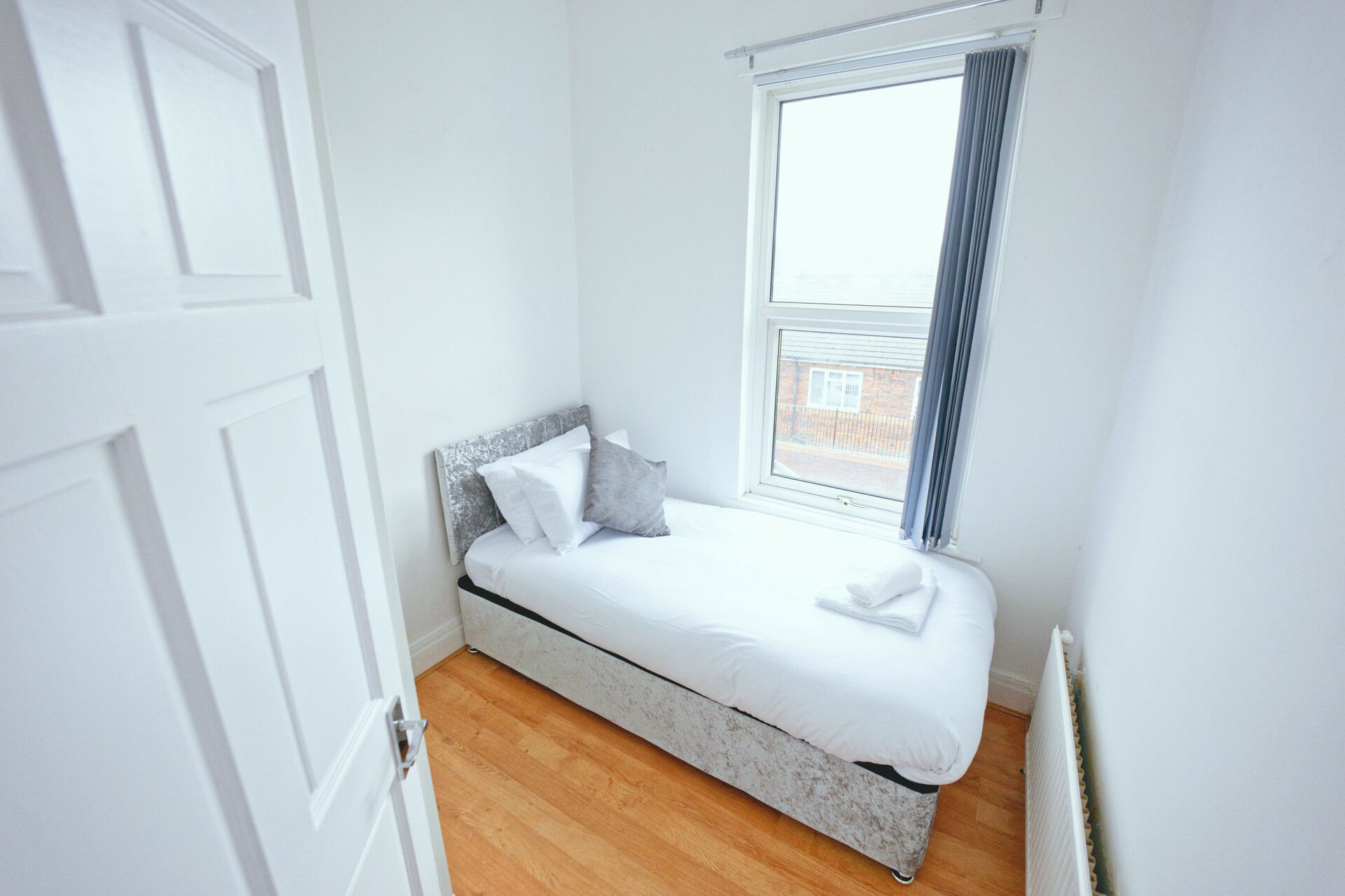 Een kleine kamer met een enkel bed, houten vloer, witte muren en een raam met gordijnen.