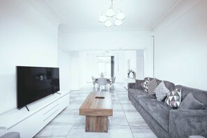 A modern living room with a flat-screen TV, a wooden coffee table, a grey sofa with patterned cushions, and a dining area in the background. - Upscale 3 BDR Modern Home in Anfield Liverpool (Liverpool)