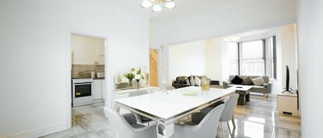 A modern kitchen with a built-in oven, a dining area with a white table and chairs, and a living room with a sofa and a TV.