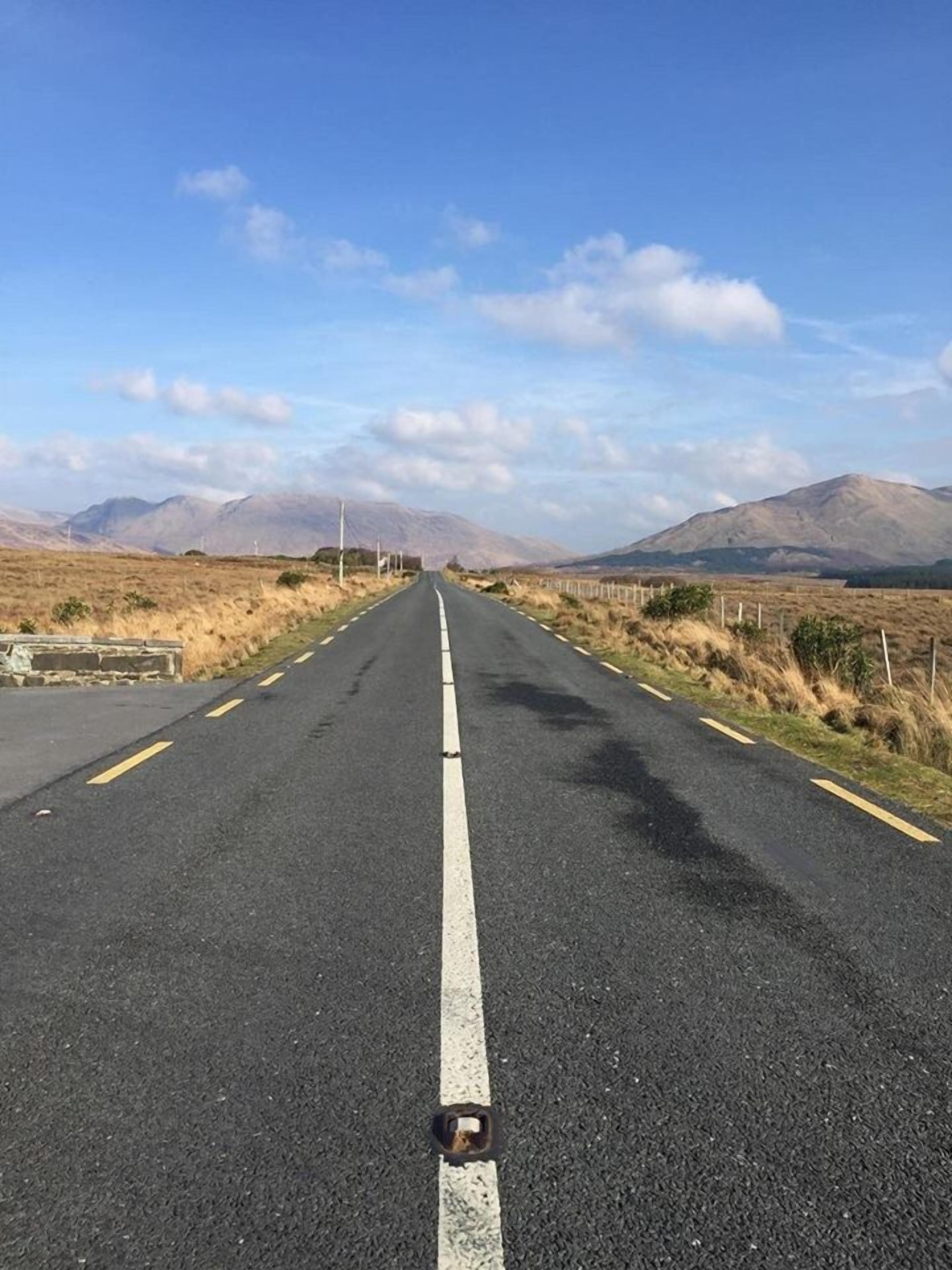 Una carretera recta con una línea blanca en el centro, bordeada por pastizales secos y montañas a lo lejos.