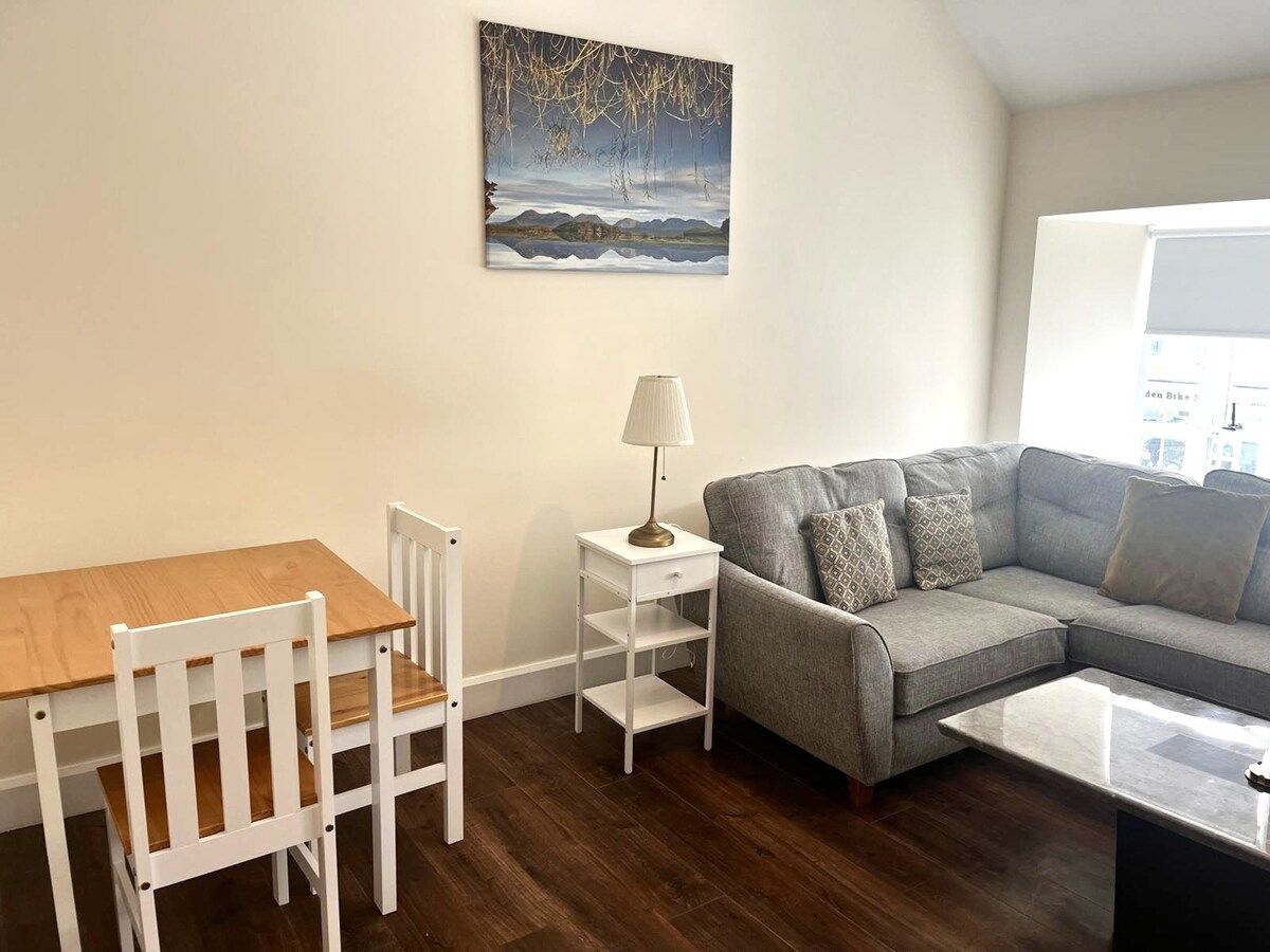 A living room with a wooden table, white chairs, a grey sofa, a white side table with a lamp, and a painting on the wall.