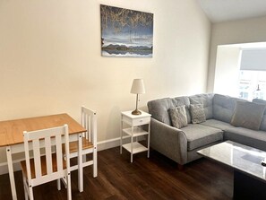 Een woonkamer met een houten tafel, witte stoelen, een grijze bank, een witte bijzettafel met een lamp, en een schilderij aan de muur.