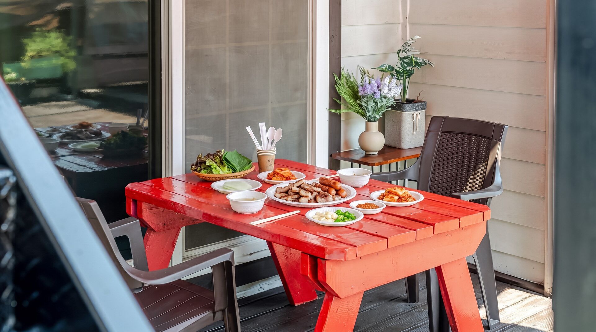 A red wooden table with various dishes, a chair, and a potted plant on a balcony.