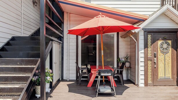 Um deck de madeira com mesa e cadeiras, um guarda-sol vermelho e uma porta decorativa.
