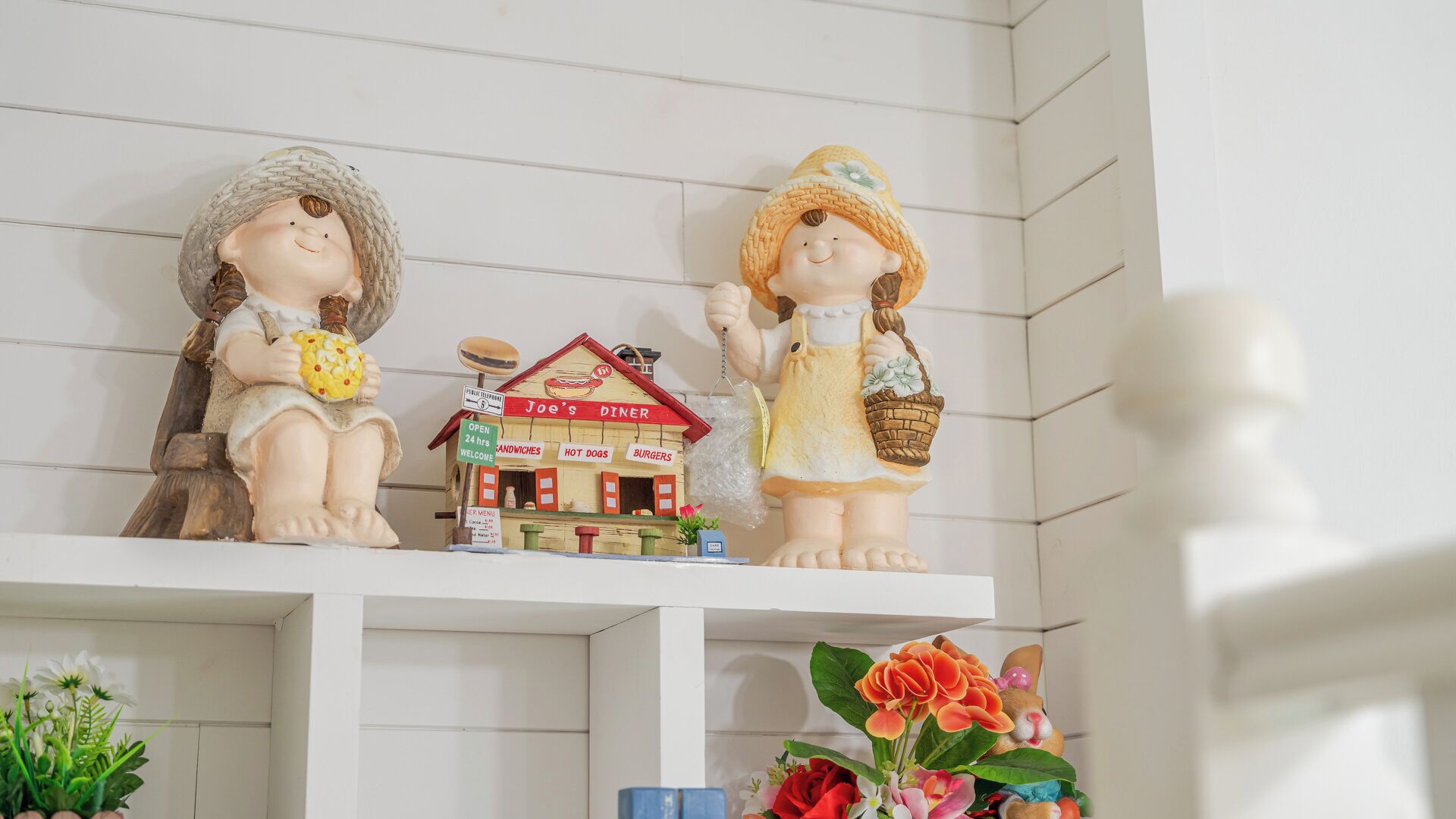 Deux statuettes sont posées sur une étagère, au-dessus d’une petite maquette de restaurant et, en dessous, un vase rempli de fleurs.