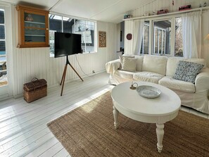 Ein Wohnzimmer mit einem weißen Sofa, einem Fernseher auf einem Dreifußständer, einer Korbo und einem runden Couchtisch.