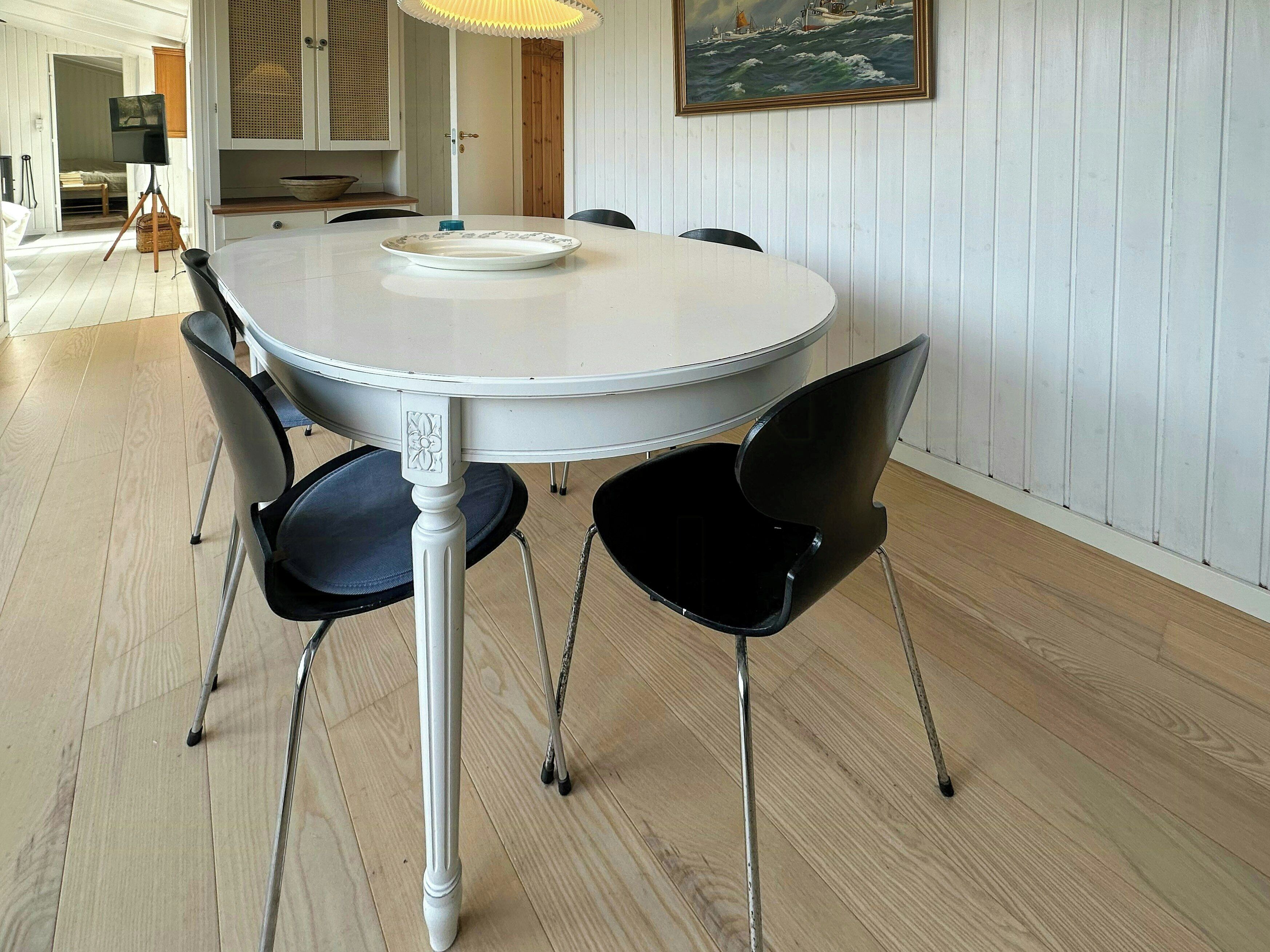Une table de salle à manger ronde et blanche, avec quatre chaises noires, un pied central blanc et un mur blanc orné d’un tableau encadré.