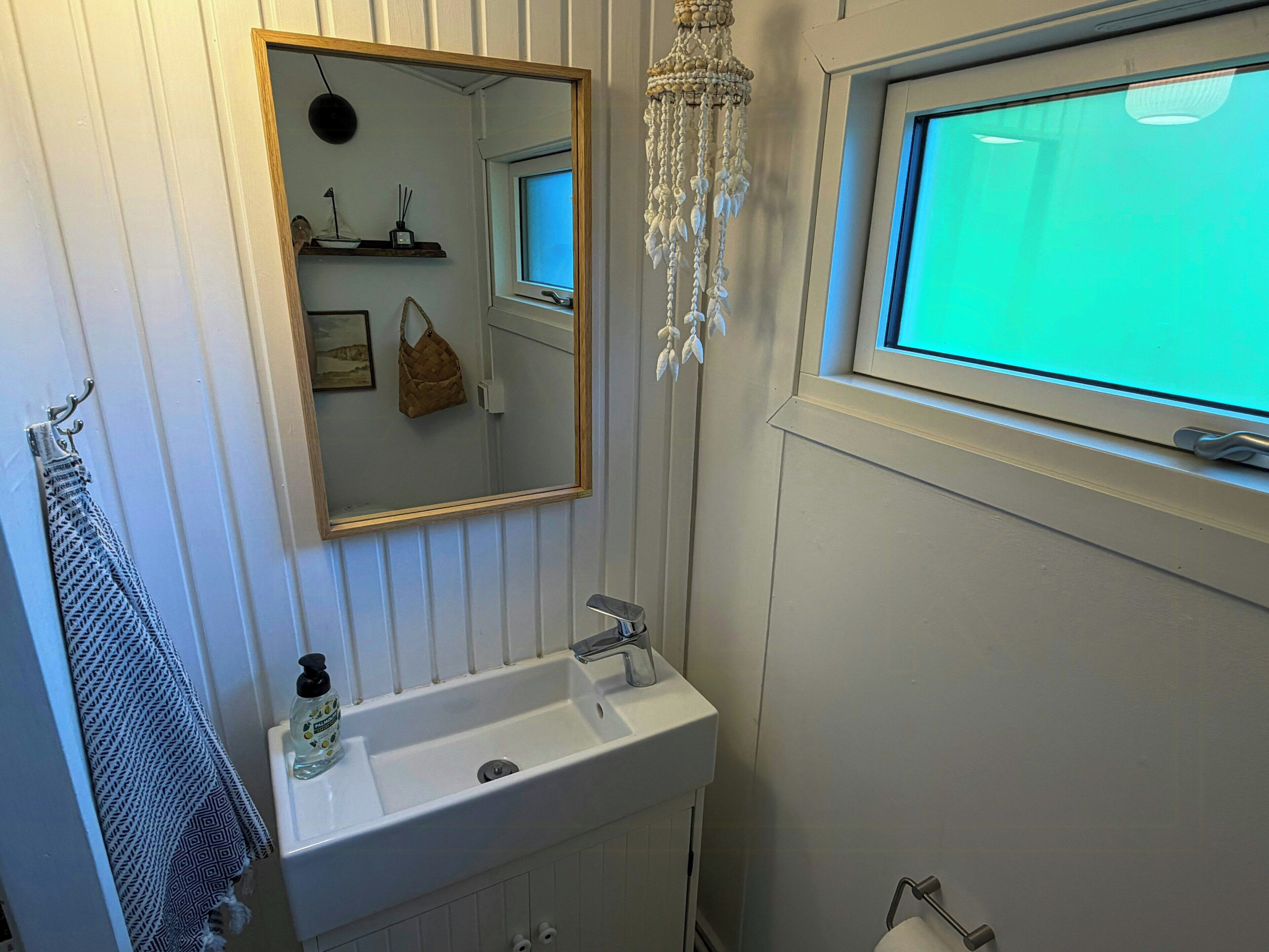 Un baño con un lavamanos blanco, un espejo, una lámpara de araña colgante, una ventana y un perchero para toallas.