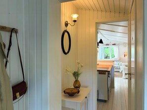 Ein Flur mit Holzboden, einer weißen Wand mit Spiegel, einem kleinen Tisch mit Blumenvase und einer Tür, die in einen Raum mit Fenster führt.