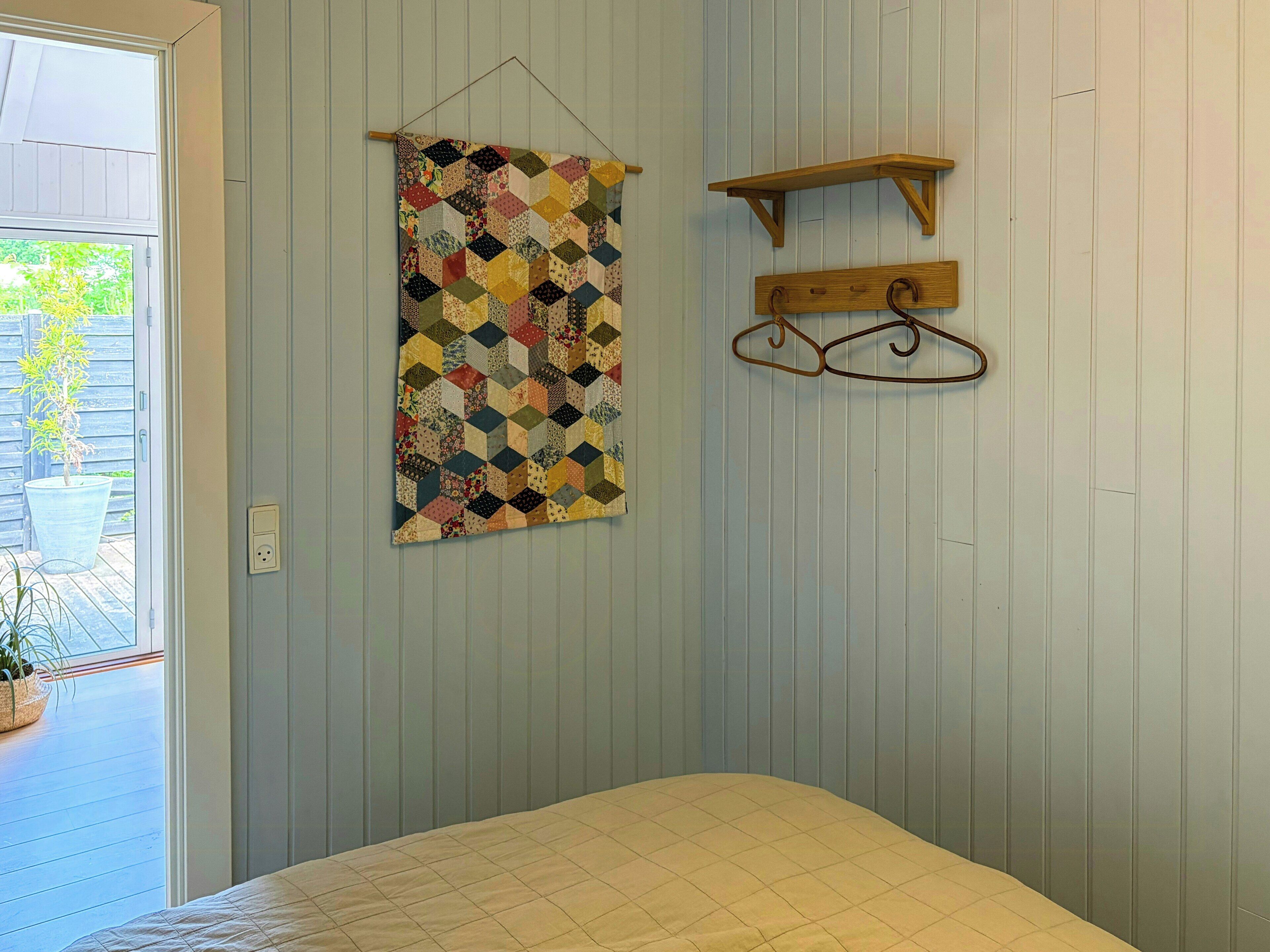 A bedroom with a bed, a hanging quilt, a wooden shelf, and a door leading to a balcony with a potted plant.